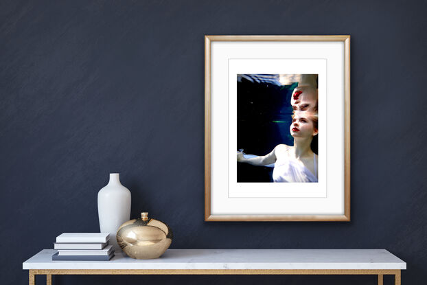 A surreal underwater scene where a girl in a white dress, who has light skin and blonde hair, is partially submerged in water and looks upon her reflection.