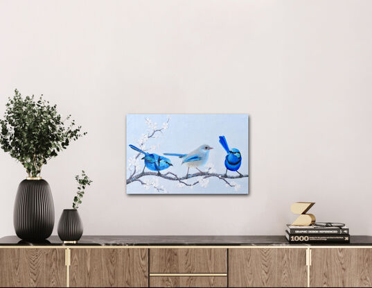 Textured oil painting of three little Splendid Fairy Wrens on a perch of white plum blossom, a male on either side of the female in the centre.  The males have a light blue head, darker blue front and back and a black stripe across the chest and near the eyes. The female is a paler grey with blue tips on the wings and tail.