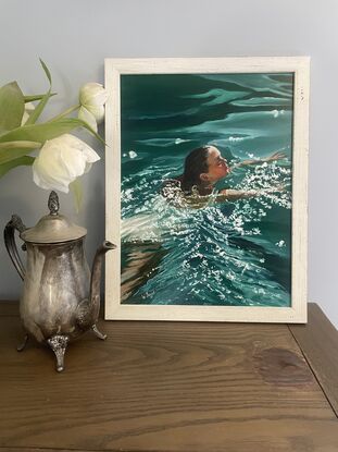 A beautiful woman with dark hair swims in a deep green ocean, sunlight dances on the water around her. Framed and ready to hang in lined white wood.