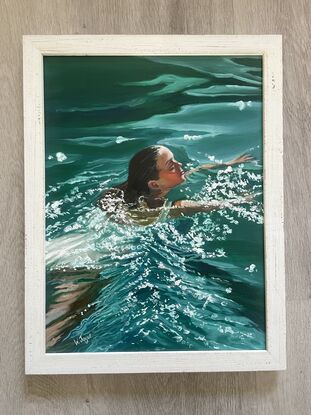 A beautiful woman with dark hair swims in a deep green ocean, sunlight dances on the water around her. Framed and ready to hang in lined white wood.