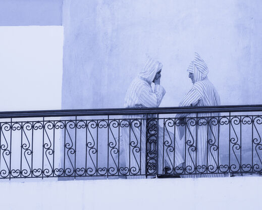 Two men on a balcony in a Moroccan city wearing tradition clothing, having a discussion.