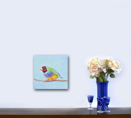 A thickly textured oil painting of a colourful Gouldian Finch, an Australian native bird. The background is a sky blue.  The little bird has a red head, green feathers on its back and purple and yellow on its front.