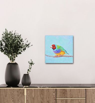 A thickly textured oil painting of a colourful Gouldian Finch, an Australian native bird. The background is a sky blue.  The little bird has a red head, green feathers on its back and purple and yellow on its front.