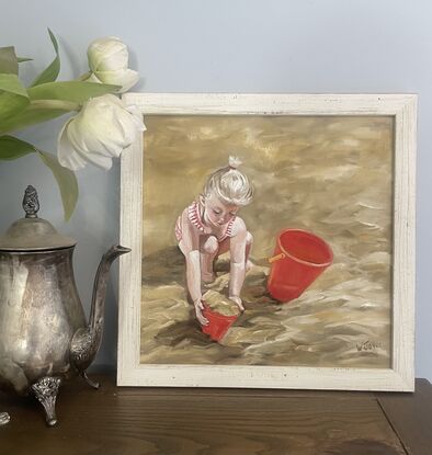 A young blued girl in red and white striped bathers crouches in golden sand playing with two red buckets. Framed in lined white wood.