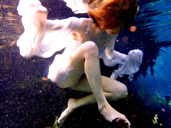 Young woman underwater showing reflections of her white dress in the swirling water whilst swimming.