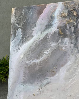 This artwork features an abstract composition with a mix of muted tones, predominantly soft grays, whites, and subtle hints of pink and mauve. There are patches of gold leaf that add a touch of metallic texture and depth to the piece. The overall effect is reminiscent of a dreamy, ethereal landscape, possibly evoking the feel of swirling clouds or a misty, mountainous terrain. 

