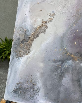 This artwork features an abstract composition with a mix of muted tones, predominantly soft grays, whites, and subtle hints of pink and mauve. There are patches of gold leaf that add a touch of metallic texture and depth to the piece. The overall effect is reminiscent of a dreamy, ethereal landscape, possibly evoking the feel of swirling clouds or a misty, mountainous terrain. 

