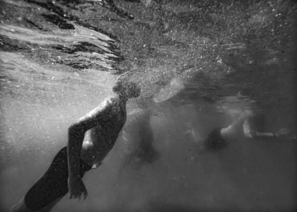 A black and white photograph of a swimmer rising up from underwater to break the surface at Merewther Ocean Baths (Newcastle). Two other swimmers are just visible in the background.  The sun through saltwater catches turbulent bubbles as the swimmer rises. 