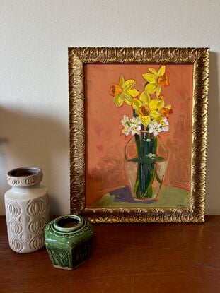 A clear rounded tall glass vase with butter yellow daffodil stesm mixed in the cream white jonquis and green grey strappy foliage. The background is ocre and the frame is gilt art nouveau in style