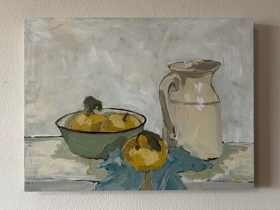 A still life scene with light green enamel bowl with light yellow nashi pears in it. To the right is an old ceramic cream coloured milk jug and they both sit on a light blue tea towel crumpled underneath. The background is white