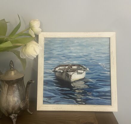 A white rowing boat with wooden trim floats front on on rippling blue water, creating an interesting reflection on the water’s surface. Framed in limed white wood and ready to hang.