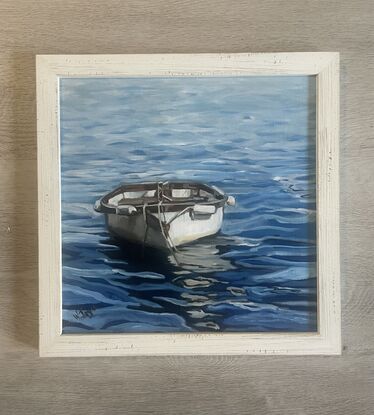 A white rowing boat with wooden trim floats front on on rippling blue water, creating an interesting reflection on the water’s surface. Framed in limed white wood and ready to hang.