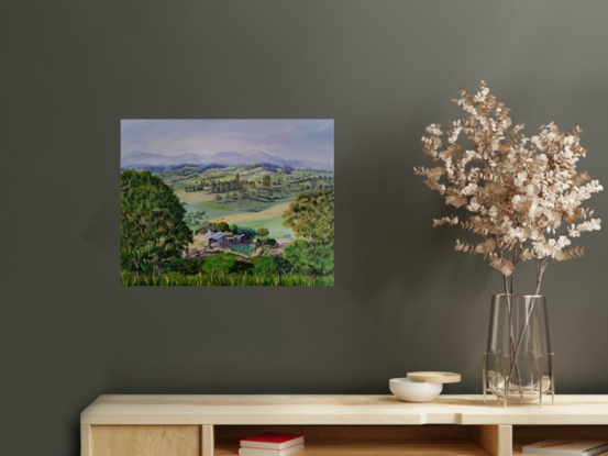 a panoramic view to the misty mountain ranges in the distance and the rolling hills of Bega Valley, with homes scattered around, surrounded by grasses, shrubs and trees with a working farm in the foreground and tall grasses and a hint of a fence in front, all framed by two large gum trees on either side of the artwork