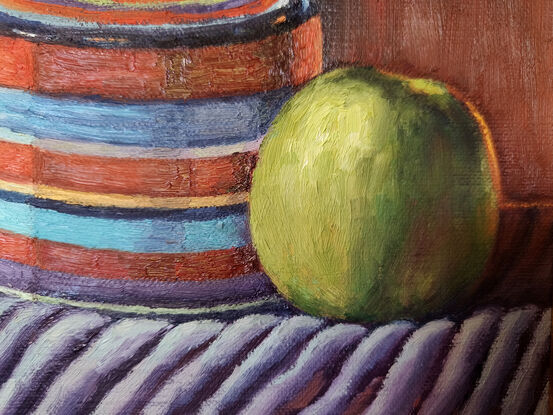 Green apple contrasted against an orange jar on  a striped fabric with light falling on them from the left direction 