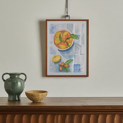 This artwork is a still life painting of lemons and cumquats in a bowl. The artist has used a variety of watercolor techniques to create a realistic and textured depiction of the fruits. The lemons are bright yellow and the cumquats are a vibrant orange. The background is a simple blue and white tile pattern which provides a contrast to the colourful fruits. The overall effect is one of freshness and abundance.