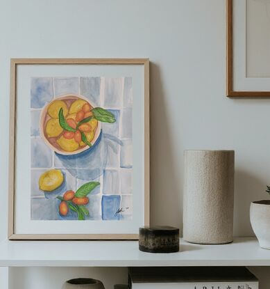 This artwork is a still life painting of lemons and cumquats in a bowl. The artist has used a variety of watercolor techniques to create a realistic and textured depiction of the fruits. The lemons are bright yellow and the cumquats are a vibrant orange. The background is a simple blue and white tile pattern which provides a contrast to the colourful fruits. The overall effect is one of freshness and abundance.