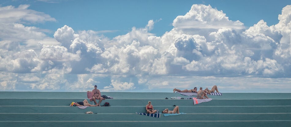 An image of Newcastle Ocean Baths. The aqua blue steps of the baths surrounds display visitors in various repose - sunbaking, sitting, sleeping in the sun. Above this, a spectacular white and purple-blue cloudscape provides a dramatic backdrop, as moisture builds from the heat of the day against brilliant blue skies. 