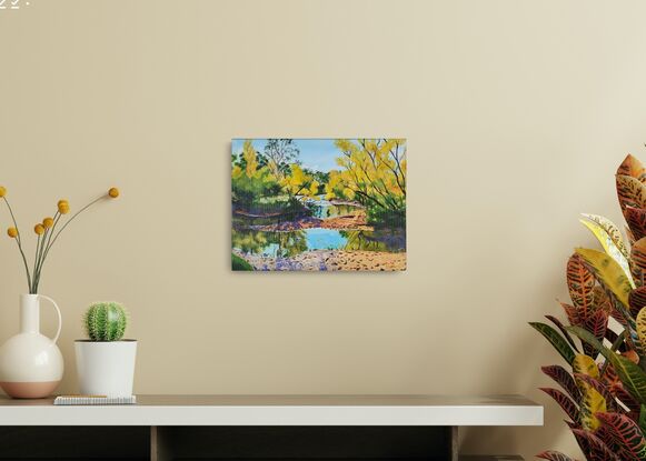 under a blue sky with some white clouds is the winding Tumut River with a number of deciduous trees glowing with cheerful colours in bright sunshine, growing along it's banks and their reflections in the slow moving water.