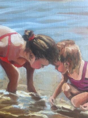 Two young girls crouch heads together in the shallows  of  the tidal break looking at something below the water’s surface.