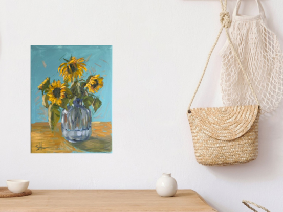 A painting of sunflowers in a clear glass vase. The sunflowers have bright yellow petals and dark brown centers, with green leaves surrounding them. The brushstrokes are loose and expressive, giving the flowers a sense of movement. The background is a soft blue, and the table is a warm, light brown. The glass vase reflects light with subtle, soft blue and gray tones, adding depth and texture to the painting. The overall feel is bright, cheerful, and inviting.