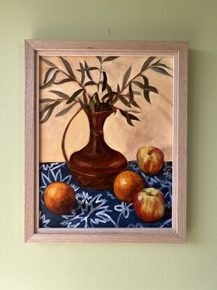 A copper jug filled with sprigs of olive leaves surrounded by oranges and apples on a blue and white patterned tablecloth. 