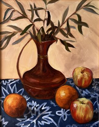 A copper jug filled with sprigs of olive leaves surrounded by oranges and apples on a blue and white patterned tablecloth. 