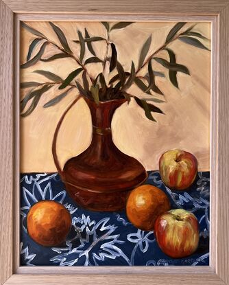 A copper jug filled with sprigs of olive leaves surrounded by oranges and apples on a blue and white patterned tablecloth. 