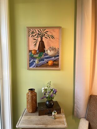 A table top with a brown vase full of olive leaves, a white decorative vintage teapot, some old mystery novels, a mustard jar and oranges on a purple striped table cloth. 