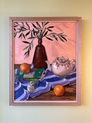 A table top with a brown vase full of olive leaves, a white decorative vintage teapot, some old mystery novels, a mustard jar and oranges on a purple striped table cloth. 