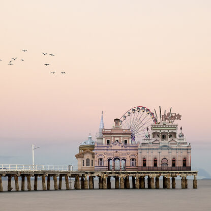 A small complex of pavilion style architecture stands at the end of a pier with a semi-hidden ferris wheel  and other rides. The background is a reverse sunset of subtle pinks with a foreground of ocean between the pavilions and the shore.