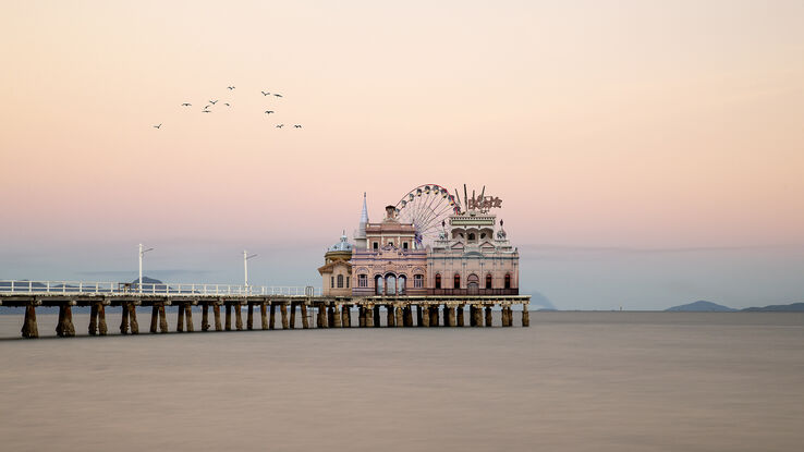 A small complex of pavilion style architecture stands at the end of a pier with a semi-hidden ferris wheel  and other rides. The background is a reverse sunset of subtle pinks with a foreground of ocean between the pavilions and the shore.