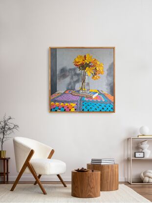 A vase of golden yellow flowers in a clear glass vase which is place on a book titled 'The Fabric of Civilization'. These are placed on a brightly coloured granny square blanket that covers the tabletop.