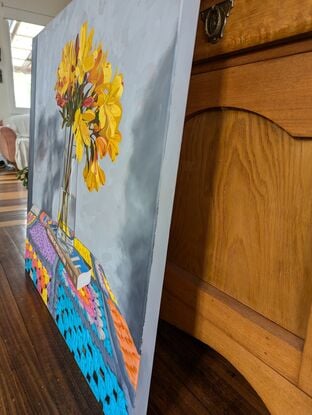 A vase of golden yellow flowers in a clear glass vase which is place on a book titled 'The Fabric of Civilization'. These are placed on a brightly coloured granny square blanket that covers the tabletop.