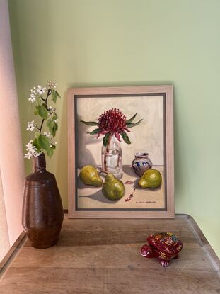 A red Waratah in a glass bottle surrounded by three pears and a small vase with blue and pink details