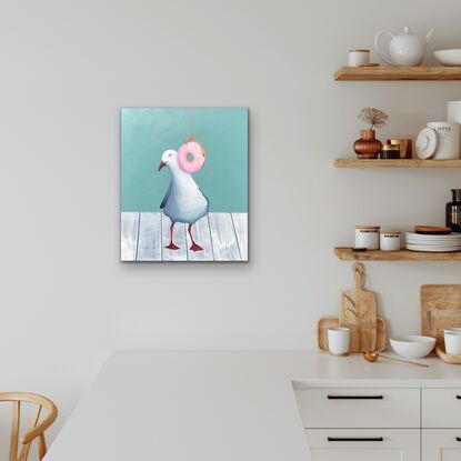 Seagull standing on a white wooden table top behind a hanging pink  donut with coloured sprinkles. 