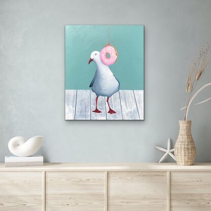 Seagull standing on a white wooden table top behind a hanging pink  donut with coloured sprinkles. 