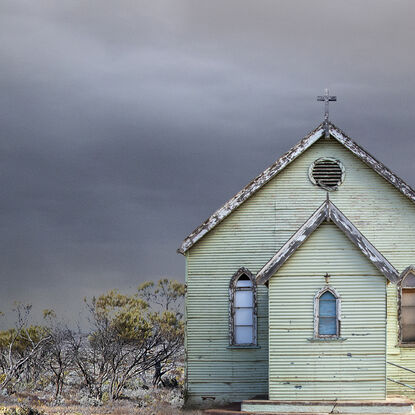 An old  retro green  weatherboard church sits alone in the Australian outback amongst some low bushes,  and a small mob of emus. The sky is very dark and stormy in the background. 