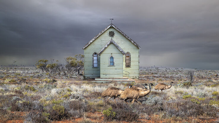 An old  retro green  weatherboard church sits alone in the Australian outback amongst some low bushes,  and a small mob of emus. The sky is very dark and stormy in the background. 