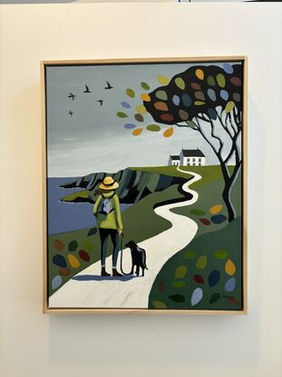 A lady wearing a green jacket, blue jeans and a yellow hat is walking her black dog along the coast along a winding white road or path that leads to a white Irish cottage. The lady had a brown long hair being blown to the side in the wind. The dog is on a leash. On the side of the white road there is green countryside with on the right a big black tree with branches with large coloured large oversized leaves. There are also some of these on the ground on each side of the path. The sky is overcast with some black birds flying. The coast has tall dark cliffs and the sea is a mud dark blue with white peaks of sea water where it meets the coastline. 