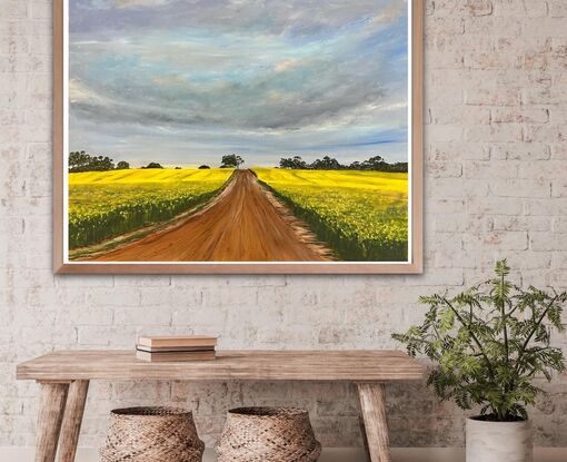 Gold field of canola with a red dirt road leading into a row of trees at the horizon 