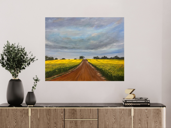 Gold field of canola with a red dirt road leading into a row of trees at the horizon 