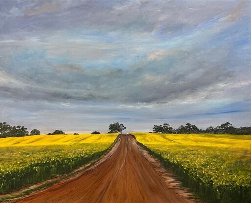 Gold field of canola with a red dirt road leading into a row of trees at the horizon 