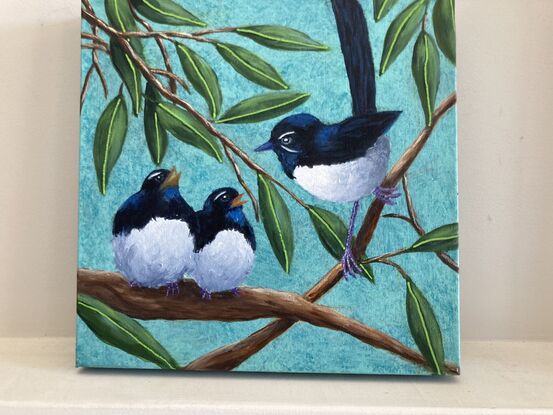 This painting is of a parent Willy Wagtail with two young chicks. The birds are black and white on a green background with branches and leaves hanging down.