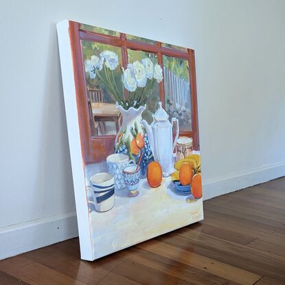 A vase with ranunculus, fruit, and crockery on a table, with a yard in the background.