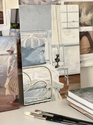 A white wrought iron bed in a bedroom with a french blue pillowcase on the pillow and brass lamp and a book on a white chair beside the bed - with sunlight pouring in the window flowing over the bed