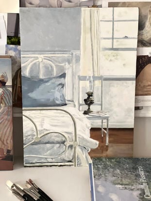 A white wrought iron bed in a bedroom with a french blue pillowcase on the pillow and brass lamp and a book on a white chair beside the bed - with sunlight pouring in the window flowing over the bed