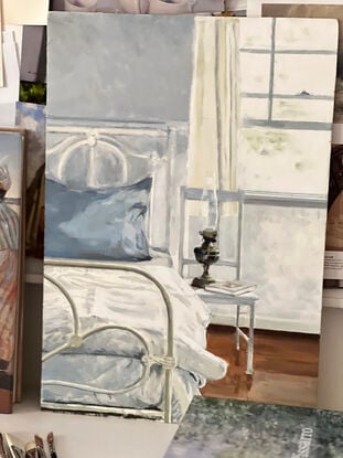 A white wrought iron bed in a bedroom with a french blue pillowcase on the pillow and brass lamp and a book on a white chair beside the bed - with sunlight pouring in the window flowing over the bed