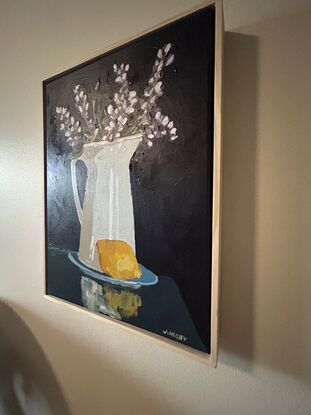 A white rustic jug serves as a vase for purple lupin flowers . it sits on a blue saucer and a lemon is in the foreground. The background is a deep blue