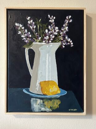 A white rustic jug serves as a vase for purple lupin flowers . it sits on a blue saucer and a lemon is in the foreground. The background is a deep blue