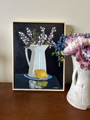 A white rustic jug serves as a vase for purple lupin flowers . it sits on a blue saucer and a lemon is in the foreground. The background is a deep blue